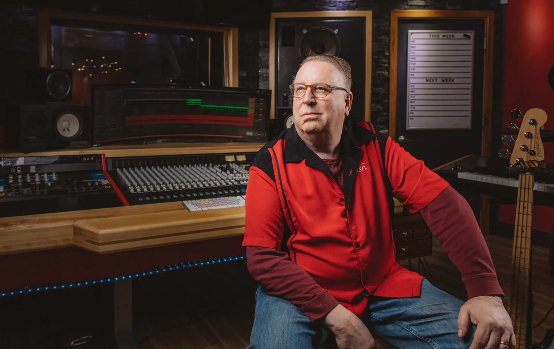 Man wearing glasses and a red shirt sitting in a recording studio with a mixing console in the background.