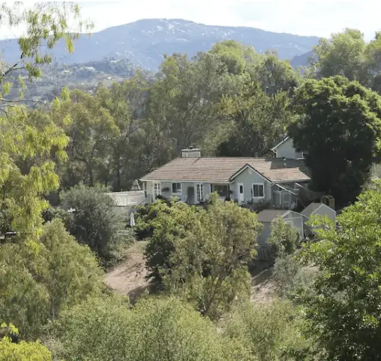 A residential house situated among trees and greenery with mountains in the background.