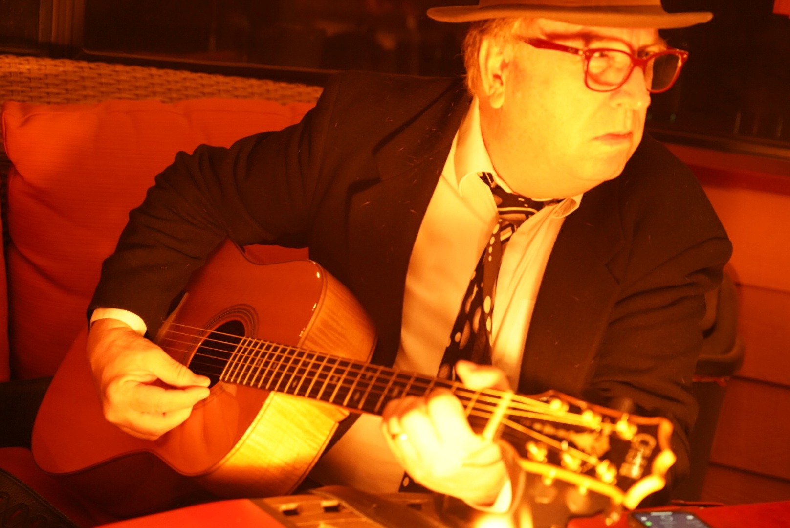 A man wearing a hat and glasses playing an acoustic guitar in a dimly lit setting.