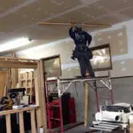 Construction worker standing on scaffolding while installing ceiling panels in a workshop.