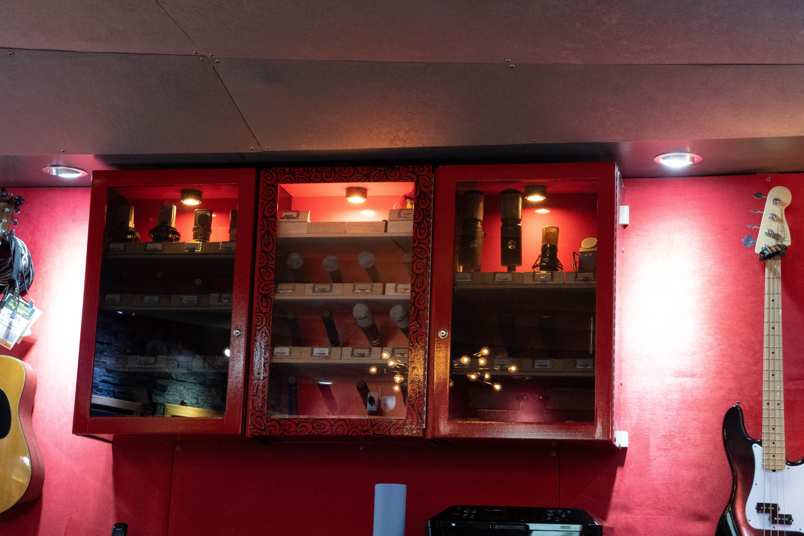 Red display cabinet containing various microphones, illuminated from above, with a guitar visible on the left.