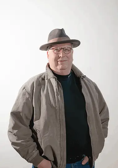 A man wearing a brown hat, glasses, and a gray jacket with a black shirt underneath, standing against a plain background.
